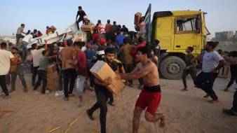L'image montre une scène animée où de nombreuses personnes semblent s'affairer autour d'un camion jaune. Des individus portent des boîtes et se déplacent rapidement, certains courant, tandis que d'autres semblent aider à décharger le camion. L'environnement est désertique, avec du sable au sol. L'atmosphère est chargée d'activité et d'urgence, indiquant qu'ils sont probablement en train de distribuer de l'aide ou des provisions.