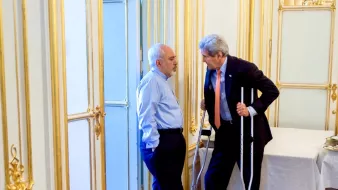 L'image montre deux hommes en train de discuter dans un environnement intérieur. L'un des hommes porte une chemise bleue et semble écouter attentivement, tandis que l'autre, utilisant des béquilles, parle avec animation. L'arrière-plan est décoré avec des moulures dorées et il y a une table couverte d'une nappe blanche. L'atmosphère semble informative et sérieuse.