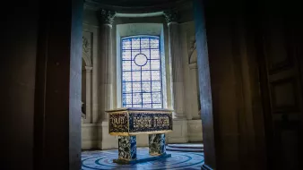 L'image montre une pièce intérieure d'un bâtiment majestueux, probablement un palais ou un musée. Au centre, il y a un grand meuble ou une caisse ornée de motifs décoratifs dorés et peints. Les murs sont en pierre, avec de très grandes fenêtres, à travers lesquelles entre une lumière douce, créant une atmosphère solennelle. Les colonnes et le sol richement décoré ajoutent à la grandeur de l'espace.