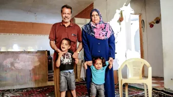 L'image montre une famille pose ensemble dans une pièce chaleureuse. Un homme et une femme, avec deux enfants, se tiennent debout sur un tapis. La femme porte un hijab et une longue robe, tandis que les enfants semblent joyeux. En arrière-plan, on peut voir des murs et une cuisine simple. L'atmosphère semble conviviale et familiale.