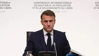 L'image montre un homme assis à une table lors d'une conférence. Il est habillé en costume sombre et porte une cravate. L'arrière-plan présente des inscriptions en français, indiquant qu'il s'agit d'une conférence internationale soutenant la population et la souveraineté du Liban, à Paris en octobre 2024. L'homme semble concentré et sérieux, regardant vers l'avant. Sur la table, on peut voir un verre d'eau et des documents.