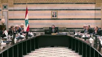 L'image montre une réunion dans une salle de conférence. Au centre, une grande table ovale est entourée de plusieurs personnes vêtues de costumes et cravates. À l'arrière-plan, on peut voir un drapeau libanais et un portrait, probablement du président du Liban, accroché au mur. Les participants semblent être à l'écoute et engagés dans la discussion, avec des documents et des ordinateurs portables devant eux. L'ambiance semble formelle et professionnelle.
