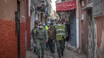 L'image montre une ruelle étroite et animée, typique d'une ville marocaine. Au premier plan, des agents de police ou des membres de la sécurité patrouillent. Ils portent des uniformes et des masques, indiquant une attention à la sécurité ou à la santé publique. En arrière-plan, on peut voir des habitants et d'autres personnes dans la rue, ainsi que des bâtiments aux façades colorées. L'atmosphère semble à la fois dynamique et surveillée, reflétant la vie quotidienne dans ce type d'environnement urbain.