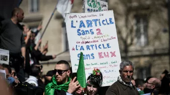 L'image montre une manifestation avec des participants tenant des pancartes. L'une des pancartes est bien visible et contient du texte en français, notamment une référence à l'article 2 et des demandes concernant cet article. Les manifestants portent des vêtements aux couleurs de l'Algérie, ce qui suggère un lien avec des préoccupations politiques ou sociales. L'atmosphère semble sereine mais déterminée, avec des gens rassemblés pour exprimer leurs revendications.