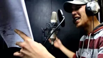 L'image montre un homme en train de chanter ou de rapper dans un studio d'enregistrement. Il porte un chapeau et des écouteurs. Devant lui, un microphone est connecté et il tient des feuilles de papier, probablement des paroles ou un texte. L'expression de son visage indique qu'il est très impliqué dans sa performance. L'arrière-plan semble sombre, ce qui est typique des environnements d'enregistrement.