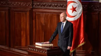 L'image montre un homme en costume, debout devant un pupitre dans un environnement officiel. Il semble être en train de prononcer un discours ou de s'adresser à un public. En arrière-plan, on aperçoit un drapeau tunisien, qui est rouge avec un cercle blanc et une étoile à cinq branches. L'atmosphère est formelle, suggérant un moment significatif, possiblement une session parlementaire ou une allocution officielle.