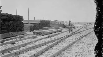 L'image montre une scène industrielle avec des voies de chemin de fer se dirigeant vers l'horizon. Sur le côté gauche, il y a des tas de planches de bois empilées, ce qui suggère une activité de construction ou de stockage. Un homme, probablement un travailleur, marche le long des rails. En arrière-plan, on aperçoit des bâtiments, donnant l'impression d'un site en pleine activité. Le ciel est dégagé, et la lumière est vive, typique d'une journée ensoleillée. L'atmosphère générale évoque une époque passée, avec une ambiance de travail acharné.