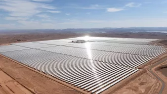 L'image montre une vaste installation de panneaux solaires, s'étendant sur une grande surface de terrain désertique. Les panneaux sont disposés en rangées régulières, reflétant la lumière du soleil. On peut apercevoir des collines à l'arrière-plan, et le ciel est clair avec quelques nuages. L'ensemble donne une impression de modernité et de durabilité, soulignant l'importance des énergies renouvelables dans le paysage contemporain.
