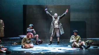 L'image montre une scène de théâtre sur une scène dépouillée. Au centre, un homme en manteau se tient avec les bras levés, tenant une bouteille dans chaque main, ce qui suggère une attitude triomphante ou exubérante. Autour de lui, plusieurs autres personnages sont assis ou accroupis, vêtus de costumes similaires. L'un d'eux semble concentré sur une activité au sol. La scène peut évoquer une ambiance dramatique ou absurde, avec des éléments de désordre comme de la poudre dispersée sur le sol. Les expressions des personnages varient, ajoutant à la dynamique de l'ensemble.