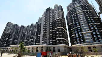 L'image montre un ensemble de grands immeubles résidentiels modernes. Les bâtiments sont principalement de couleur noire et blanche, avec de grandes fenêtres qui laissent entrer la lumière. On peut voir un chantier en cours au premier plan, avec des véhicules de construction et des matériaux dispersés. Il y a aussi quelques arbres et des espaces verts, suggérant un aménagement paysager en développement. Le ciel est dégagé et ensoleillé.