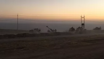L'image montre un paysage de chantier au crépuscule, avec un ciel teinté de couleurs douces allant du jaune à l'orange. On aperçoit des machines de construction et des travailleurs en silhouettes, entourés de poussière. À l'arrière-plan, on distingue des poteaux électriques. Le sol semble sec et rocailleux, suggérant une zone de travaux en cours. L'atmosphère est calme, mais il y a une impression d'activité et d'effort en cours.
