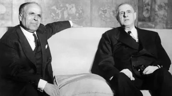 L'image montre deux hommes assis sur un canapé dans un environnement formel. Ils ont l'air sérieux et portent des costumes sombres. L'un des hommes semble légèrement se pencher en arrière, tandis que l'autre regarde devant lui. L'arrière-plan est décoré, mais les détails ne sont pas clairement visibles. L'atmosphère générale apparaît académique et solennelle.