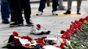 L'image montre un sol avec une paire de chaussures de sport blanches laissées à l'écart. Autour des chaussures, il y a une traînée de fleurs rouges, probablement des œillets, qui sont disposées en ligne. On peut également voir d'autres objets sur le sol, tels que des photos et des éléments décoratifs. En arrière-plan, des gens sont visibles, mais leur apparence et leurs détails ne sont pas clairement discernables. L'ensemble de la scène dégage une atmosphère de mémoire ou de commémoration.