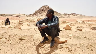 L'image montre deux personnes assises dans un paysage désertique. L'un des hommes est en train de s'asseoir sur une grosse pierre, portant une veste en cuir et des lunettes de soleil. Il semble pensif, observant le paysage autour de lui. En arrière-plan, une autre personne est également assise, mais davantage éloignée. Le terrain est constitué de sable et de roches, avec des formations rocheuses au loin, sous un ciel clair. L'atmosphère semble calme et isolée.