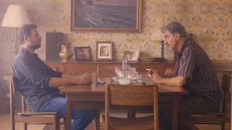 L'image montre deux hommes assis à une table en bois dans une pièce décorée avec un papier peint aux motifs rétro. L'ambiance est intérieure et intimiste, avec une lampe à côté et des photos accrochées au mur. Les deux hommes semblent engagés dans une conversation sérieuse. Le premier homme, à gauche, est vêtu d'une chemise sombre et regarde l'autre. Le deuxième homme, à droite, fume une cigarette et semble pensif. L'éclairage doux ajoute à l'atmosphère.