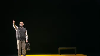 L'image montre un homme sur scène, debout sur un tapis jaune. Il porte un pull noir par-dessus une chemise blanche et tient une serviette ou un sac à main à la main. L'homme semble pointer vers le haut, exprimant une émotion forte ou un message. La scène est sombre autour de lui, ce qui met l'accent sur sa présence au centre et sur l'objet noir qui pourrait être un banc ou une table. L'ambiance donne une impression de sérieux ou de réflexion.