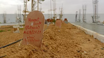 L'image montre un cimetière avec des tombes marquées par des plaques en terre cuite. Le sol est sablonneux et légèrement irrégulier, avec quelques plantes éparses. En arrière-plan, on remarque des structures en construction, probablement des bâtiments ou des monuments. Le ciel semble nuageux, créant une atmosphère calme mais mélancolique. Les inscriptions sur les plaques indiquent les noms et des dates, suggérant que ces êtres chers ont été récemment enterrés.