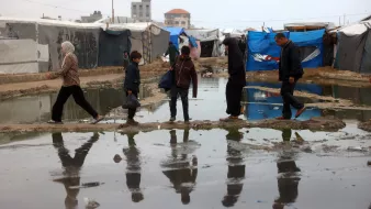 L'image montre des personnes marchant à travers un camp de tentes, avec des surfaces détrempées et des flaques d'eau. On peut voir une femme en hijab, un enfant, et plusieurs adultes, tous avançant sur des passages surélevés ou des morceaux de terrain au-dessus de l'eau. L'environnement semble difficile, et les refuges sont constitués de bâches et de tissus. Les reflets des marcheurs se voient dans les flaques, ajoutant une dimension visuelle à la scène.