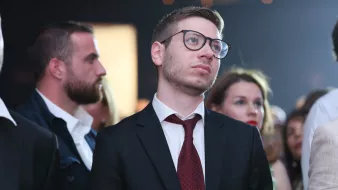 L'image montre un groupe de personnes rassemblées lors d'un événement. Au premier plan, un homme porte un costume noir avec une chemise blanche et une cravate marron. Il a des lunettes et semble pensif, le regard tourné vers le haut. À l'arrière-plan, d'autres personnes, dont une femme et un homme, sont également présentes, ajoutant à l'atmosphère de l'événement. La lumière est douce, créant une ambiance élégante.