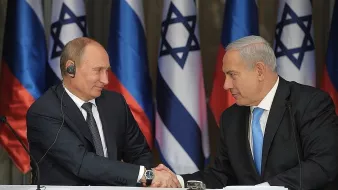 L'image montre deux hommes en costume, se serrant la main, dans un cadre formel. À leur arrière-plan, on peut voir des drapeaux de la Russie et d'Israël. L'un des hommes, à gauche, semble être en train de porter un écouteur, tandis que l'autre, à droite, regarde attentivement. L'ambiance suggère un moment de coopération ou d'accord.