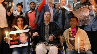 L'image montre un groupe de personnes rassemblées, tenant des bougies allumées. Au premier plan, un homme en fauteuil roulant est au centre, tenant une pancarte. Les autres personnes autour de lui semblent participer à une manifestation pacifique, certaines brandissant des affiches et d'autres affichant des expressions sérieuses. L'ambiance semblerait être axée sur la défense des droits et de la liberté d'expression.