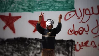 L'image montre un jeune garçon, les yeux bandés, levant les bras en signe de victoire ou de protestation. Il se trouve devant un mur sur lequel sont inscrits des slogans en arabe, ainsi qu'un symbole représentant le drapeau syrien. Cette scène évoque des thèmes de résistance et de luttes dans un contexte de conflit.