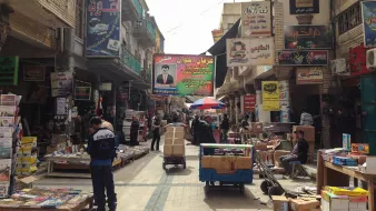 L'image montre une rue animée, typique d'un marché urbain dans une ville. On peut voir des étals et des magasins de part et d'autre, présentant divers produits, avec des affiches et des panneaux publicitaires colorés accrochés aux murs. Certaines personnes sont visibles, allant et venant, tandis que d'autres semblent être en train de travailler, probablement en déchargeant des marchandises. Le sol est souvent encombré de caisses et d'autres objets, créant une ambiance vivante et dynamique. Des parasols sont également présents, offrant un peu d'ombre dans cette rue fréquentée.