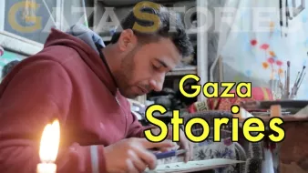 L'image montre un homme assis, probablement en train de travailler sur un projet créatif ou artisanal. Il porte un sweat à capuche rouge et semble concentré sur son activité. À côté de lui, une bougie est allumée, ce qui indique qu'il pourrait travailler dans un environnement peu éclairé. En arrière-plan, on aperçoit des éléments de décoration ou des fournitures, ce qui suggère une atmosphère artistique. En haut de l'image, on peut lire "Gaza Stories", ce qui laisse penser que le contexte est lié aux récits ou aux témoignages de Gaza.