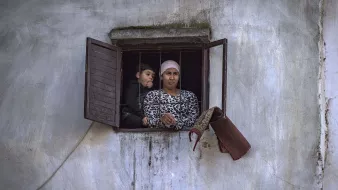 L'image montre deux personnes regardant à travers une fenêtre. Il y a une femme, vêtue d'un haut à motifs, qui semble attentive. À côté d'elle, un enfant, visiblement curieux, se penche légèrement. L'environnement est simple, avec des murs en crépi et une fenêtre aux volets ouverts. L'atmosphère semble réfléchie et calme.