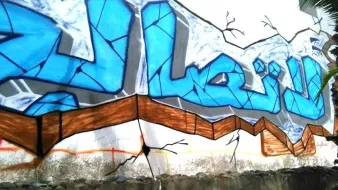 L'image montre un mur de graffiti avec des lettres stylisées en bleu. Le fond est légèrement cassé, ce qui donne l'impression que le mur est en train de se fissurer. Le mot écrit semble être en arabe, avec des motifs et des contours en noir qui accentuent le design. En bas, on peut apercevoir des déchets et de l'herbe, ce qui montre que l'art se trouve dans un environnement urbain.