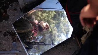 L'image montre un miroir fragmenté reposant sur le sol, reflétant l'image d'un homme en uniforme avec un fusil. À l'arrière-plan, on voit des arbres, suggérant un environnement naturel ou une zone de conflit. Le reflet donne une perspective intrigante sur la scène. L'homme semble vigilant, peut-être en attente ou en préparation pour une opération.