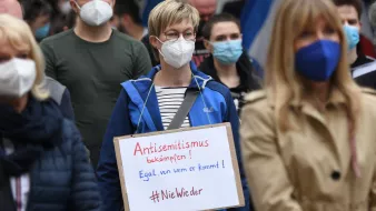L'image montre une manifestation où des personnes portent des masques. Au premier plan, une femme se distingue avec une pancarte sur laquelle il est écrit en allemand "Antisemitismus bekämpfen! Egal, von wem er kommt! #NieWieder". Elle porte une veste bleue et se tient parmi d'autres manifestants, tous masqués. L'ambiance semble sérieuse et engagée en faveur de la lutte contre l'antisémitisme.