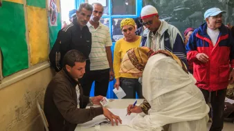 L'image montre un groupe de personnes se rassemblant autour d'un bureau dans une salle. Un homme est assis à une table, où il semble aider une femme, qui est en train de signer un document. Elle porte une tenue claire et un foulard. D'autres personnes, dont des hommes et des femmes, observent la scène, manifestant un intérêt. Les murs sont décorés de couleurs vives, et une fenêtre ouverte laisse entrer la lumière. L'environnement semble informer d'une activité communautaire, peut-être liée à un vote ou à une inscription.