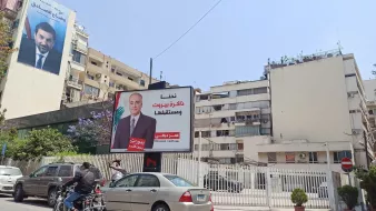 L'image montre une rue animée avec des voitures et des motos en circulation. À gauche, il y a une grande affiche sur un bâtiment, probablement une campagne politique, mettant en avant une personne, avec des textes en arabe. À côté, un autre bâtiment est visible, avec une façade claire et quelques plantes qui ajoutent une touche de verdure à l'environnement urbain. Le ciel est bleu, ce qui donne une ambiance ensoleillée à la scène.