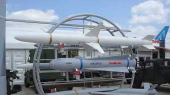 L'image montre une exposition de missiles sur un socle. On peut voir plusieurs types de missiles, dont un missile de croisière, un missile air-air et un missile guidé. Les missiles sont présentés sur un support circulaire, avec des informations en dessous de chacun d'eux. En arrière-plan, on aperçoit des véhicules militaires et un ciel partiellement nuageux. C'est une scène typique d'une foire ou d'une exposition de défense militaire.