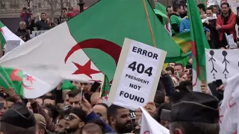 L'image montre une manifestation avec de nombreuses personnes brandissant des drapeaux algériens. Au premier plan, un manifestant tient une pancarte avec l'inscription "ERROR 404 PRESIDENT NOT FOUND", suggérant un mécontentement vis-à-vis de la situation politique. L'ambiance semble être celle d'une contestation, avec une foule réunie pour exprimer ses revendications.
