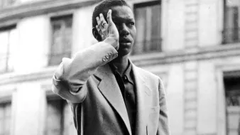 L'image montre un homme en ville, levant une main près de son front comme s'il saluait ou réfléchissait. Il porte un costume élégant qui semble bien ajusté. L'arrière-plan présente des bâtiments typiques d'une ville européenne, avec des fenêtres et des balcons. La photo est en noir et blanc, ce qui lui donne une ambiance vintage et nostalgique.