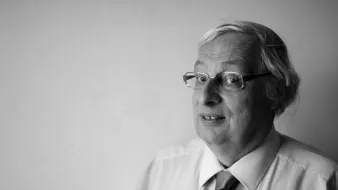 L'image montre un homme d'âge moyen avec des cheveux gris et des lunettes. Il porte une chemise claire avec une cravate. Il regarde légèrement sur le côté avec un sourire, le fond étant neutre et clair. L'image est en noir et blanc, ce qui lui donne un aspect classique et intemporel.