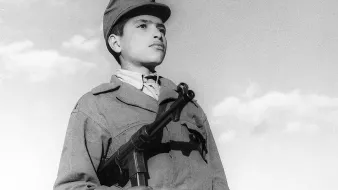 L'image montre un jeune garçon en uniforme militaire, tenant une arme à feu. Son regard est sérieux et déterminé, tandis qu'il se tient droit, face à l'objectif. Le ciel en arrière-plan est clair, avec quelques nuages. L'uniforme et l'arme peuvent indiquer qu'il fait partie d'un contexte militaire ou historique spécifique. L'ambiance de la photo évoque un sentiment de courage et de bravoure.