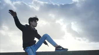 L'image montre un jeune homme assis sur un muret, avec une posture décontractée. Il porte un sweat noir et un jean bleu, avec des baskets noires. Ses cheveux sont légèrement ébouriffés par le vent, et il semble apprécier le moment en regardant vers le ciel, qui est nuageux avec quelques éclaircies. L'ambiance générale évoque une sensation de liberté et de fraîcheur.