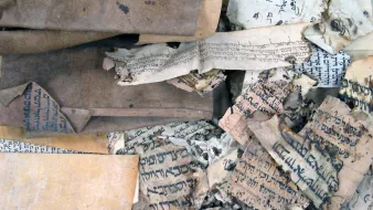 L'image montre un ensemble de vieux manuscrits et de documents enroulés et usés. Les pages sont usées, certaines sont jaunies et déchirées, et on peut voir des inscriptions écrites dans des langues anciennes et probablement des alphabets différents. La texture des papiers varie, certains étant plus épais que d'autres, et la composition générale donne une impression d'antiquité et de fragilité, suggérant leur importance historique.