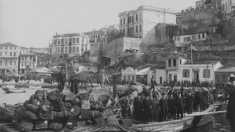 Cette image en noir et blanc montre une scène portuaire historique. On peut voir une barge chargée de marchandises, notamment des fûts et des sacs, avec des personnes debout dessus. En arrière-plan, il y a des bâtiments qui semblent faire partie d'une ville côtière, avec des structures en pierre et des maisons aux balcons. La côte est animée, avec d'autres bateaux et des gens sur le rivage, créant une atmosphère de commerce maritime. La lumière et le contraste évoquent une ambiance d'époque.