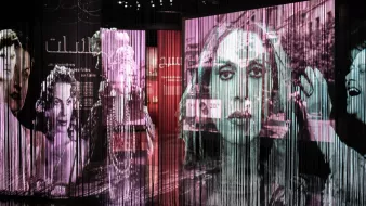 L'image présente une installation artistique composée de plusieurs grands panneaux transparents sur lesquels sont projetées des images en noir et blanc de visages féminins. Les projections créent un effet de profondeur et de mouvement, avec des formes et des silhouettes qui se superposent. Le fond est teinté de couleurs douces, suggérant une ambiance à la fois artistique et contemplative. Ce type d'installation évoque une réflexion sur la représentation et l'identité, tout en intégrant des éléments visuels captivants.