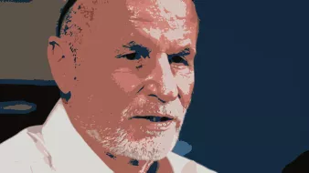 L'image représente un homme avec des cheveux grisonnants et une barbe. Il porte une chemise blanche et des lunettes posées sur sa tête. Son expression semble sérieuse et il a l'air de parler ou d'écouter attentivement. Le fond est de couleur sombre, ce qui met en valeur son visage.