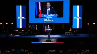 Un homme parle sur scène, avec des drapeaux français en arrière-plan.