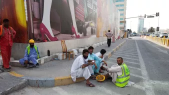 L'image montre un groupe d'hommes assis sur un trottoir, probablement en train de faire une pause. Ils portent des vêtements traditionnels, et certains portent des gilets de travail. En arrière-plan, on aperçoit un grand panneau publicitaire avec des images colorées. L'environnement urbain est visible, avec des bâtiments modernes et une route. L'atmosphère semble calme, et les hommes semblent discuter entre eux.