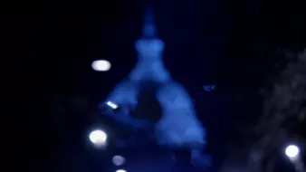 L'image montre une scène nocturne avec un éclairage diffus. Au centre, on aperçoit une silhouette floue, probablement d'un bâtiment ou d'une structure, avec un dôme et une pointe. Les lumières autour ajoutent une ambiance mystérieuse et éthérée, créant un contraste entre l'obscurité environnante et les sources lumineuses. L'atmosphère semble calme et légèrement mélancolique.