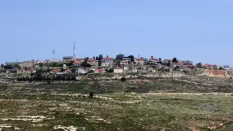 L'image montre un village situé sur une colline, avec des maisons dispersées, certaines avec des toits rouges. On peut également voir des installations comme des antennes de communication. Le paysage environnant est composé de champs et de collines vertes, suggérant une région rurale. Le ciel est clair et bleue, ce qui donne une ambiance tranquille à la scène.