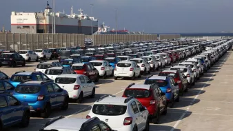 L'image montre un grand parking de voitures, organisé en rangées bien alignées. On aperçoit plusieurs types de voitures de différentes couleurs, majoritairement en blanc, rouge et bleu. En arrière-plan, on peut voir des bateaux de marchandises amarrés dans un port, sous un ciel nuageux. Cette scène semble se dérouler dans une zone portuaire, où les véhicules sont probablement en attente d'expédition ou de distribution.