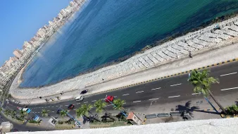 L'image montre une vue panoramique d'une côte en bord de mer. On aperçoit une route qui longe la plage, avec des palmiers sur le côté. L'eau se présente dans des teintes de bleu et de vert, et on peut voir des bâtiments en fond, probablement la ville ou des complexes résidentiels. La lumière du soleil illumine la scène, donnant une atmosphère agréable et ensoleillée.